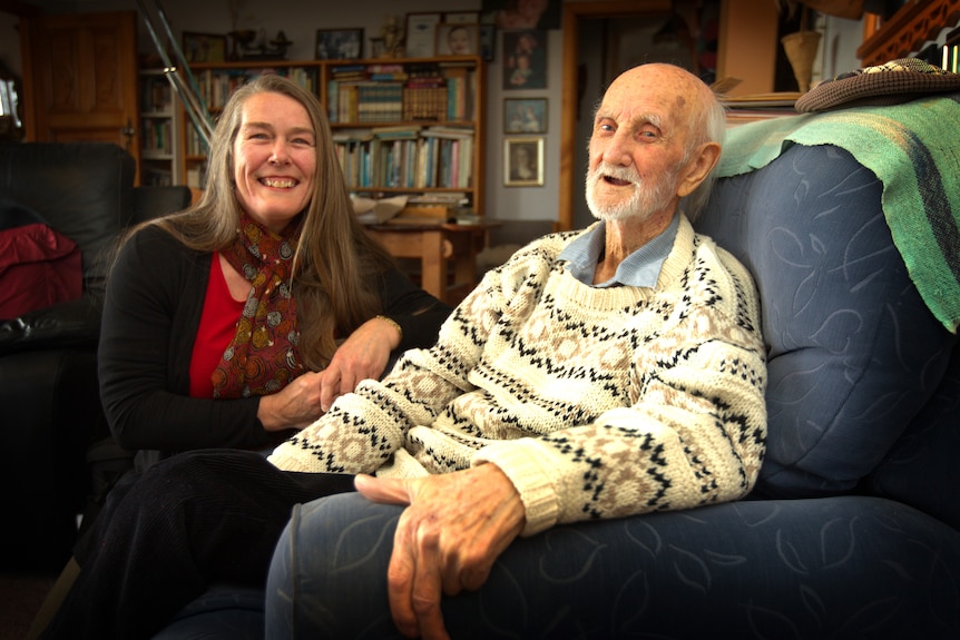 A very old man sitting in a soft chair with a woman crouched by his side, both smiling