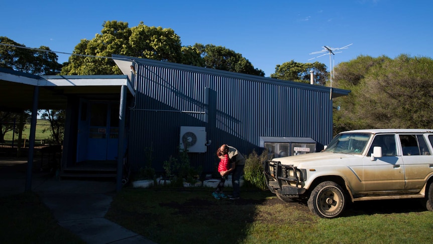 A girl wearing a red jacket hugs her father in a patch of sunlight in front of a school building and beside a dirty truck.