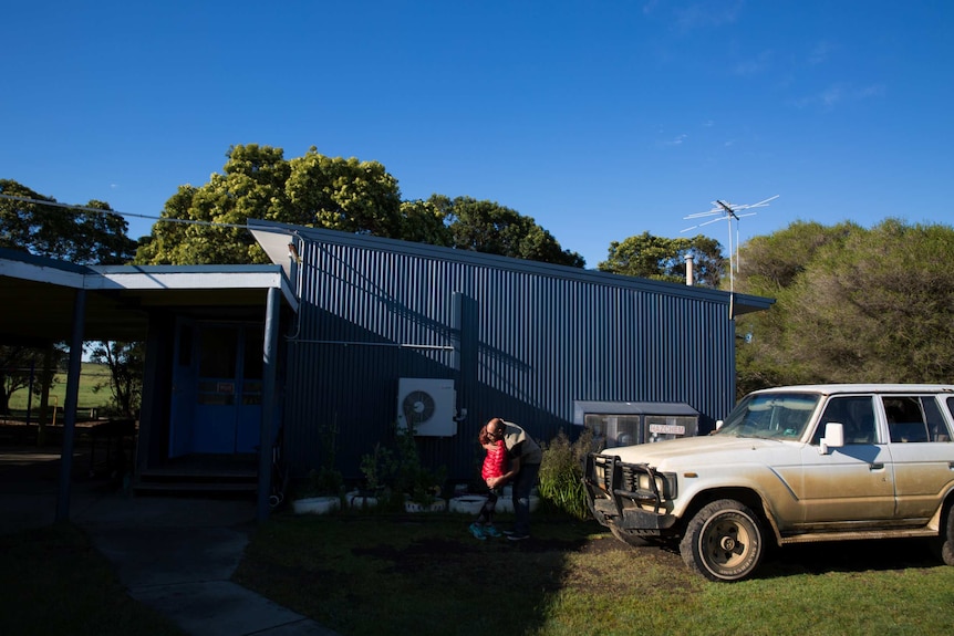 A girl wearing a red jacket hugs her father in a patch of sunlight in front of a school building and beside a dirty truck.