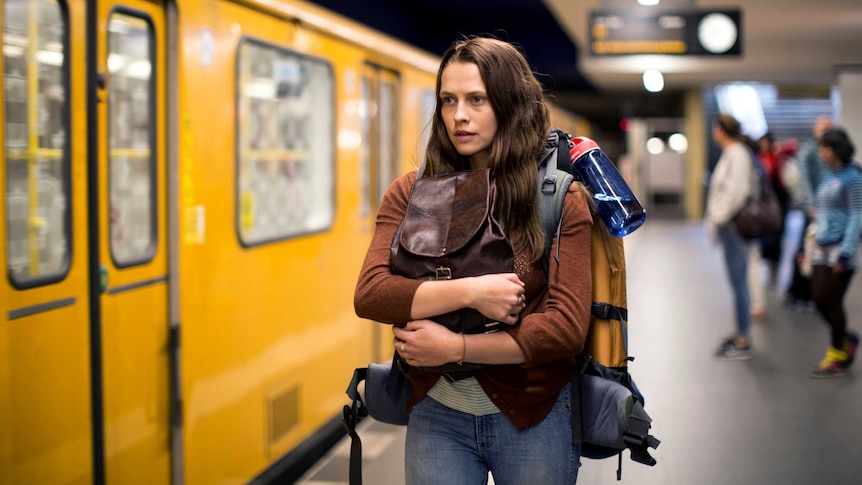 A 20-something woman with long brown hair on a train platform, holding her backpack to her chest.
