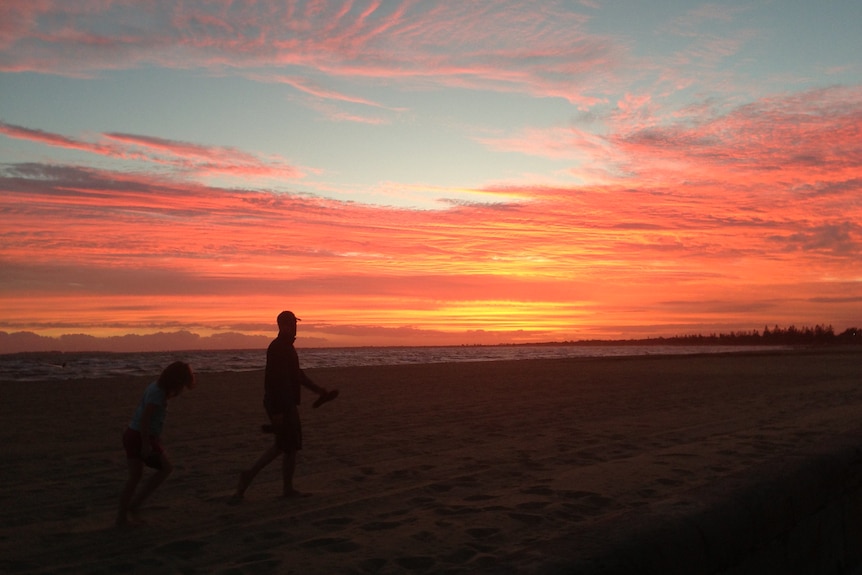 Two people walk along Altona Beach, in front of a magnificent pink and orange sunset.