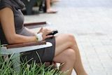 A woman sits on a bench using a mobile phone. Her face is not visible.