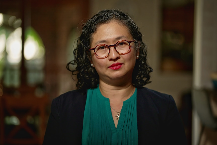 A woman with short, dark, curly hair, small rounded glasses and red lipstick wearing an emerald green blouse and black cardigan.