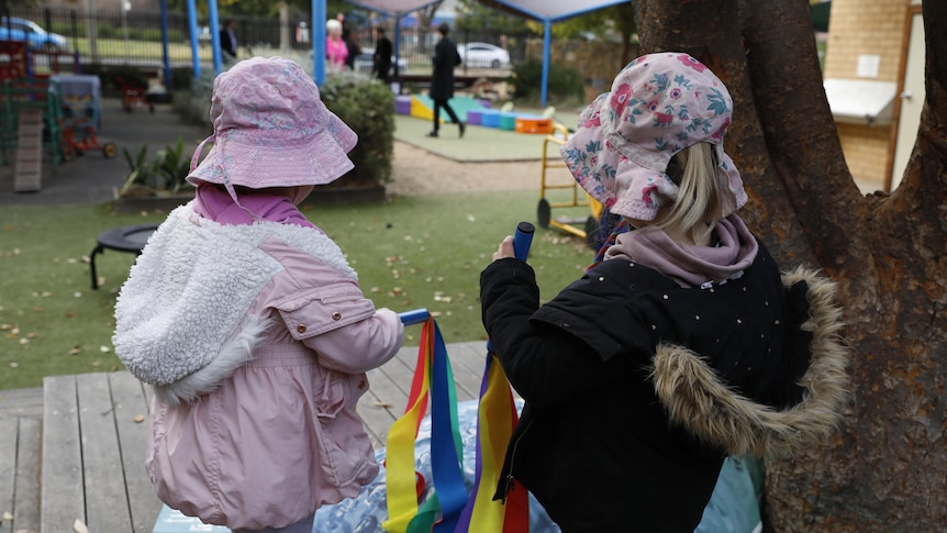 Two young girls wearing pink hats and holding rainbow streamers on an outdoor deck at a childcare centre