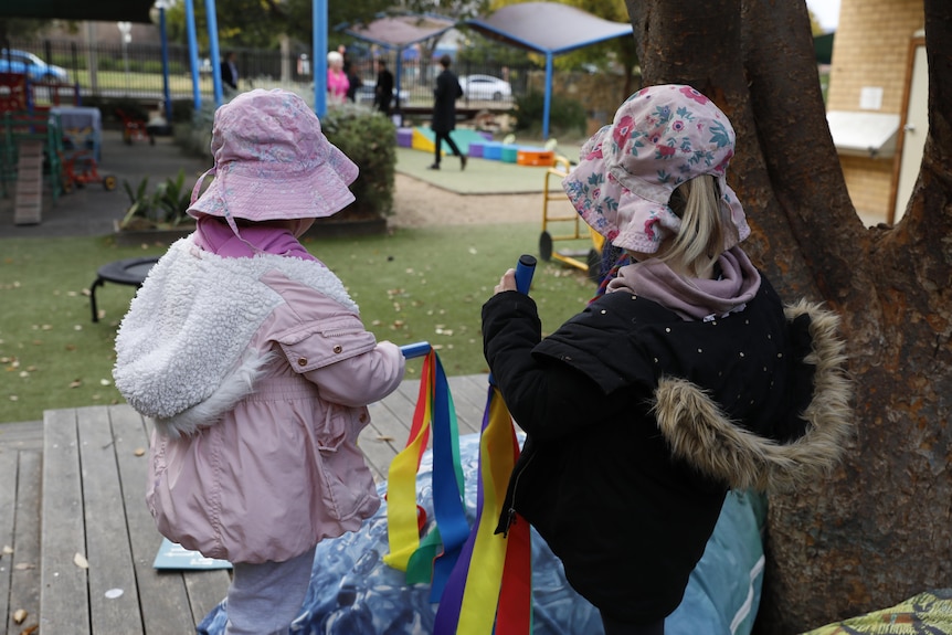 Two young girls wearing pink hats and holding rainbow streamers on an outdoor deck at a childcare centre