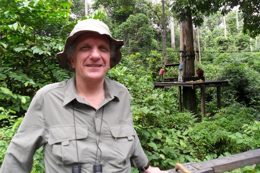 Man wearing khaki clothing and binoculars around neck is surrounded by jungle-like trees