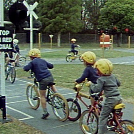 Children in yellow helmets wait with bikes at a miniature road crossing inside a traffic park.