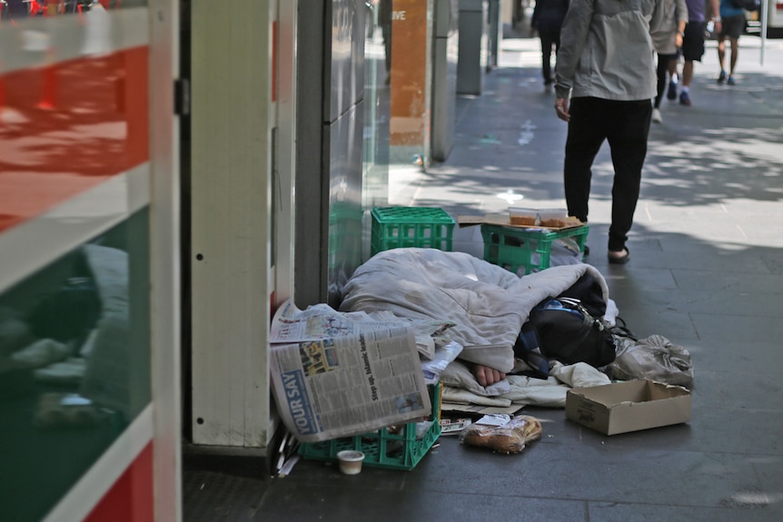 A person wrapped in a blanket, surrounded by their belongings, outside a 7-11 store.