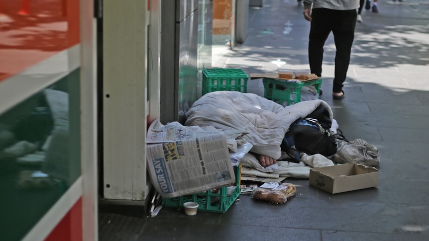 A homeless person sleeps on a street covered in blankets and surrounded by milk crates.