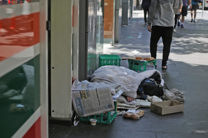 A homeless person sleeps on a street covered in blankets and surrounded by milk crates.