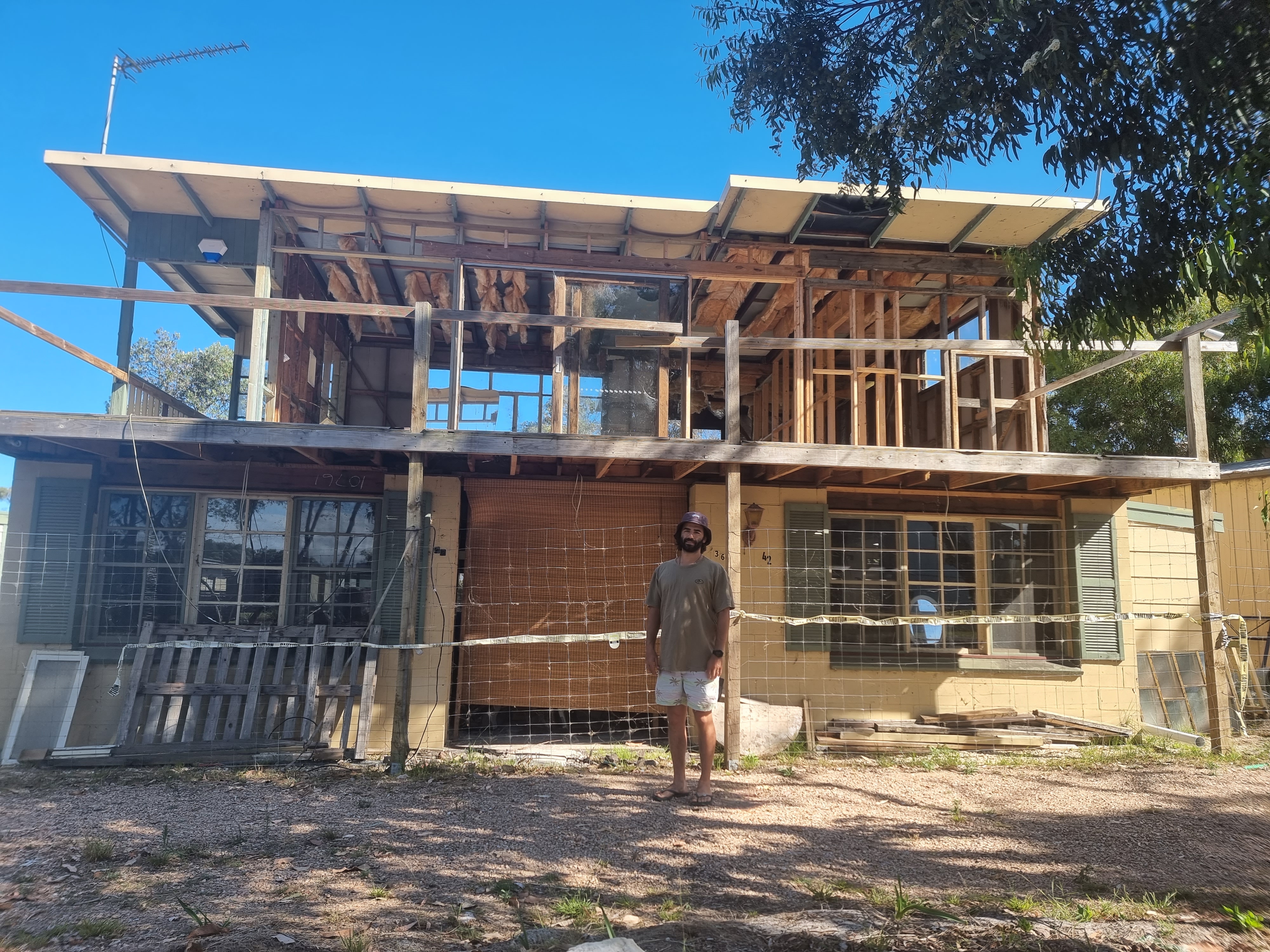 A man in shorts and tee and wearing a hat, black beard, thongs, stands in front of a half finished house.