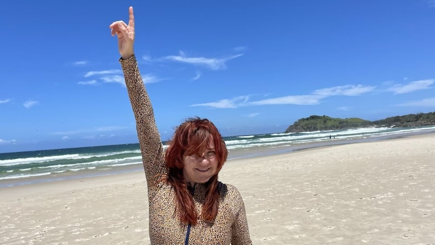 A woman with auburn hair stands on the beach with her hand in the air