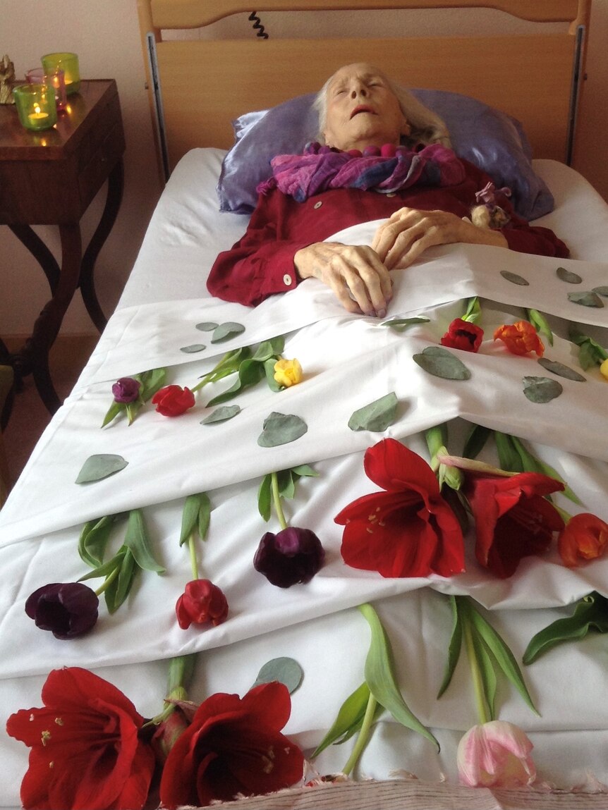 An older woman, now dead, lies on a bed surrounded by fresh flowers 