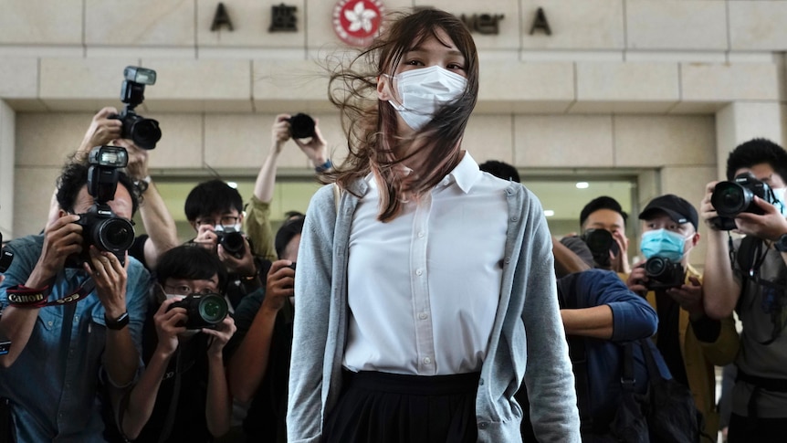 Woman wearing grey cardigan, white shirt and mask stands in front of a crowd of cameras. 