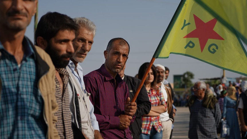 Kurdish civilians wave Kurdish flags and look unhappy as they watch US troops pass by.