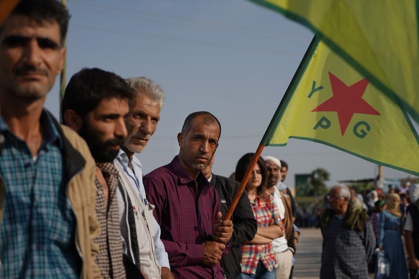 Kurdish civilians wave Kurdish flags and look unhappy as they watch US troops pass by.