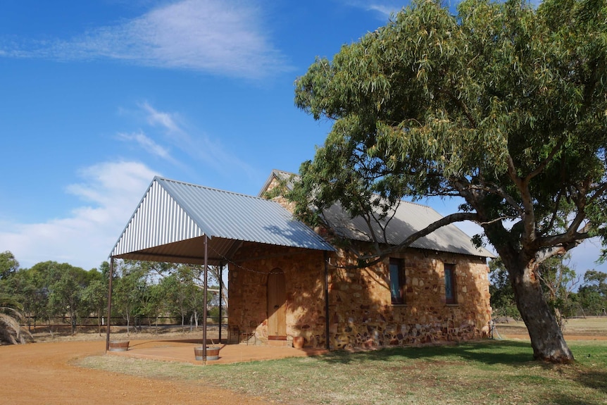 A tall tree casts a shadow on a small, old  limestone church with a corrugated roof and verandah. The sky is bright blue.