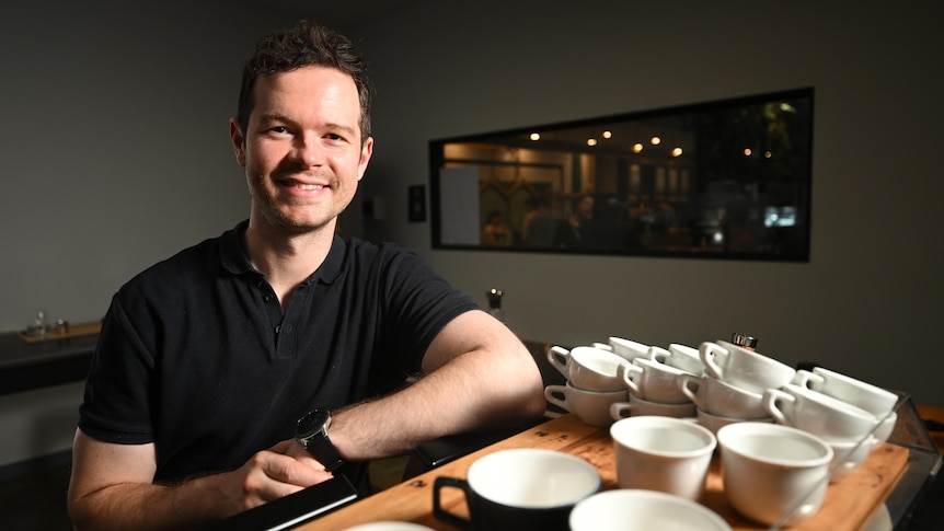 barista anthony douglas smiles and leans on a coffee machine with coffee cups stacked on it