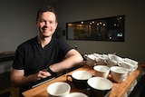 barista anthony douglas smiles and leans on a coffee machine with coffee cups stacked on it