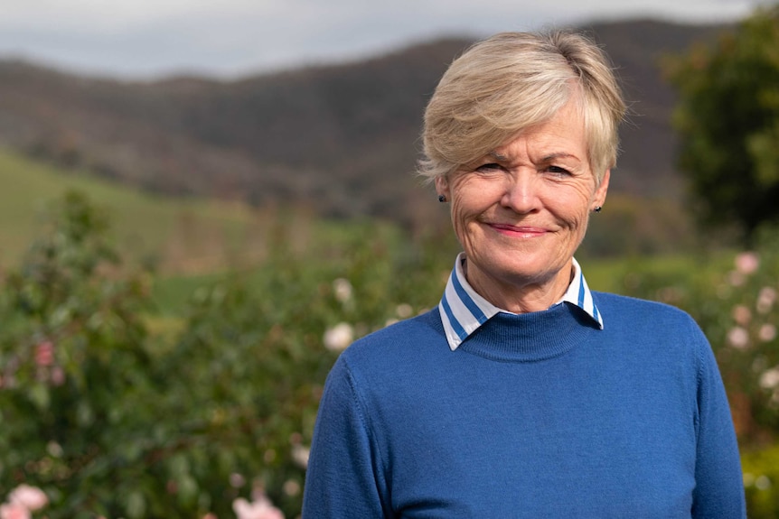A women with short blonde hair, wears a bright blue jumper and shirt. Blurred in the background is green hills and burnt hills.