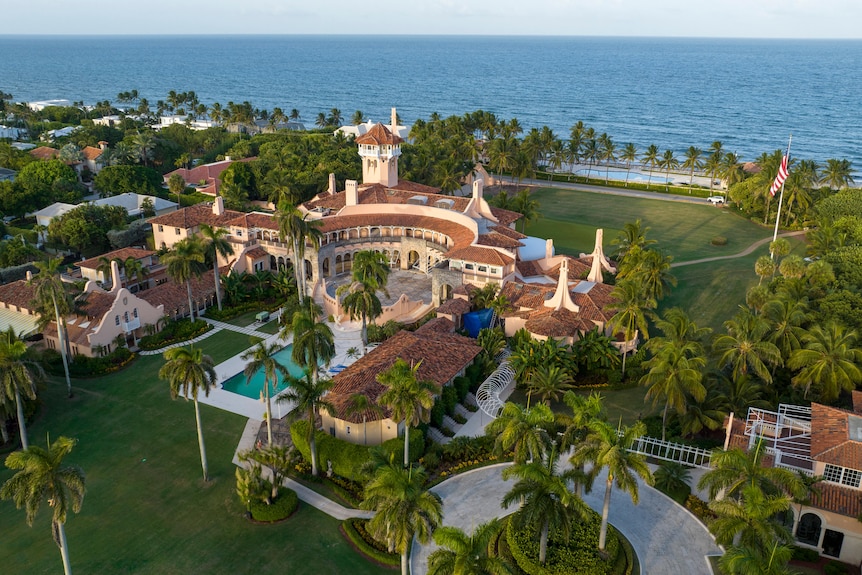 A huge mansion with palm trees and a pool is seen from above near the ocean.