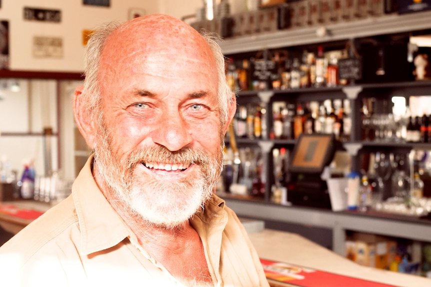 A bald man with a grey beard smiling into the camera with a public bar in the background.