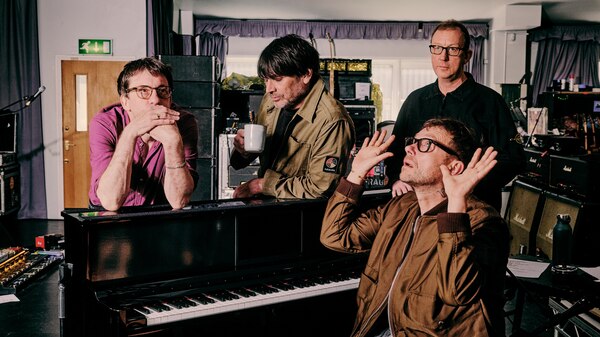 Blur's Damon Albarn sits at a piano hands comically reaching up, Graham Coxon, Alex James and Dave Rowntree stand behind