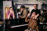 Blur's Damon Albarn sits at a piano hands comically reaching up, Graham Coxon, Alex James and Dave Rowntree stand behind 