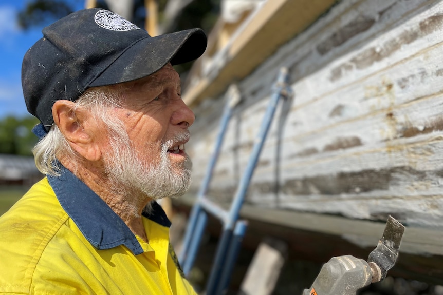 A side profile of the face of an older man with grey hair and a beard, holding a work tool next to an old boat.