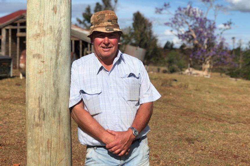 A man wearing a light blue shirt and camouflage hat leaning against a telegraph pole on a property 