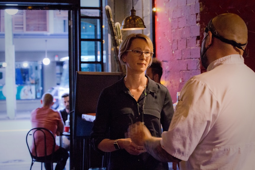 Kate Cole stands talking to JC Miranda in front of wide open doors that lead from the restaurant onto the street.