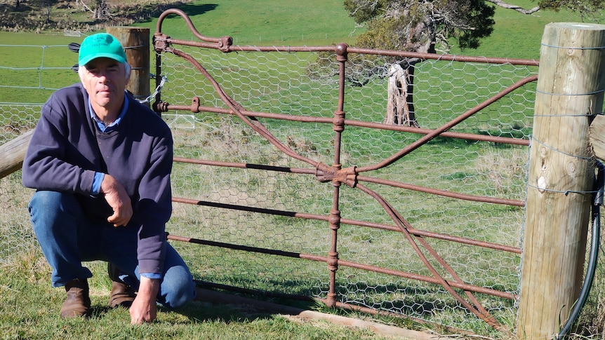 A farmer in blue jumper and green cap squats next to a rusty gate. 