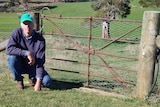 A farmer in blue jumper and green cap squats next to a rusty gate. 