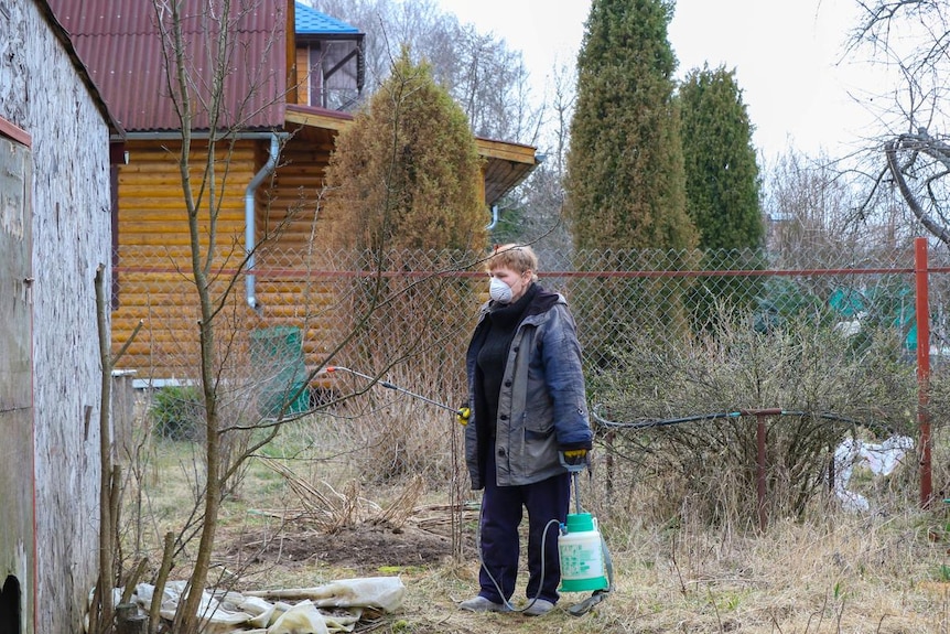 A woman wearing a mask and holding a canister stands in a yard.