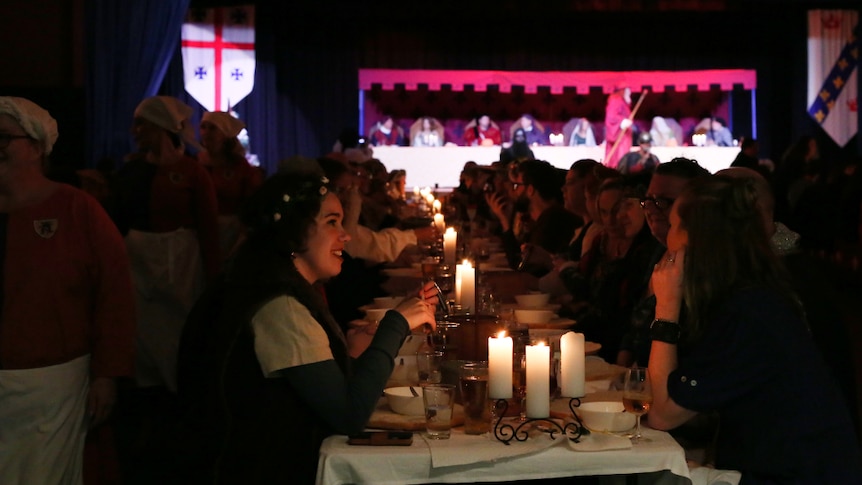 Diners in medieval dress sit at a long, candle-lit table. while servants pass behind. A high table is in the background.