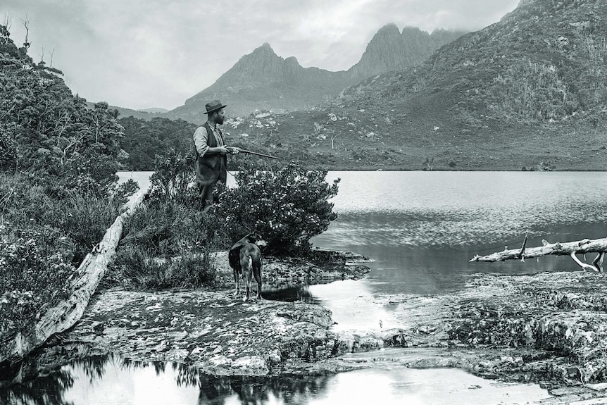 Black and white historic image of a man with a gun standing at the edge of a lake, Cradle Mountain in the background.