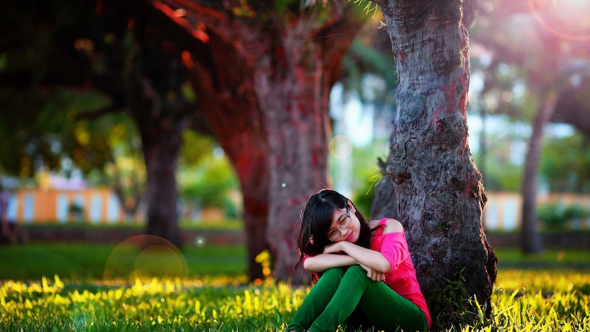 A young woman sits in the shade behind a tree in a park