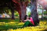 A young woman sits in the shade behind a tree in a park