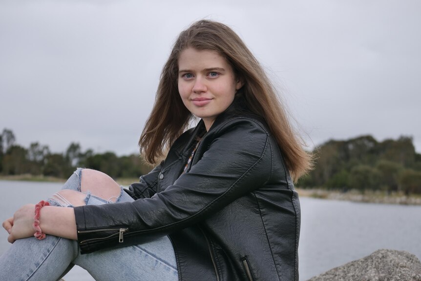 A girl wearing a black leather jacket and blue ripped jeans hugs her knees on a rock overlooking a lake.