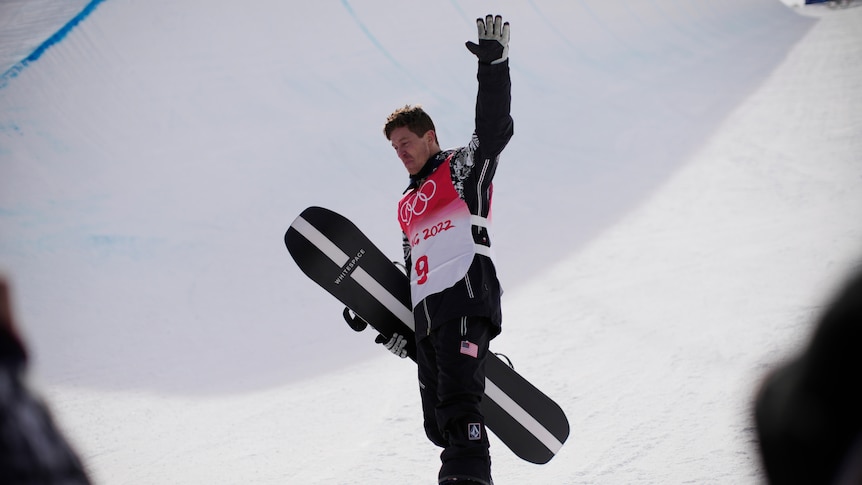 A man holding a snowboard waves.
