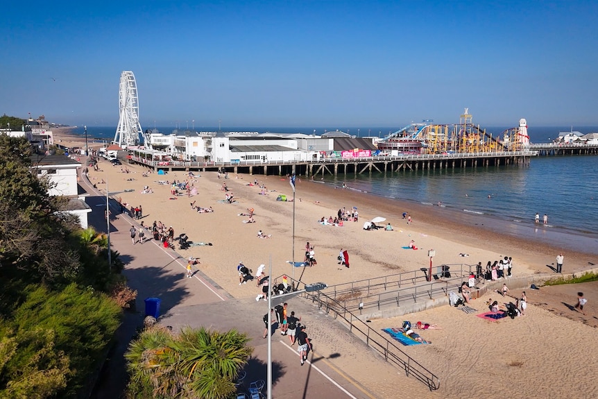 Beachgoers are scattered over the sand on a beach, with a pier and amusement park in the distance