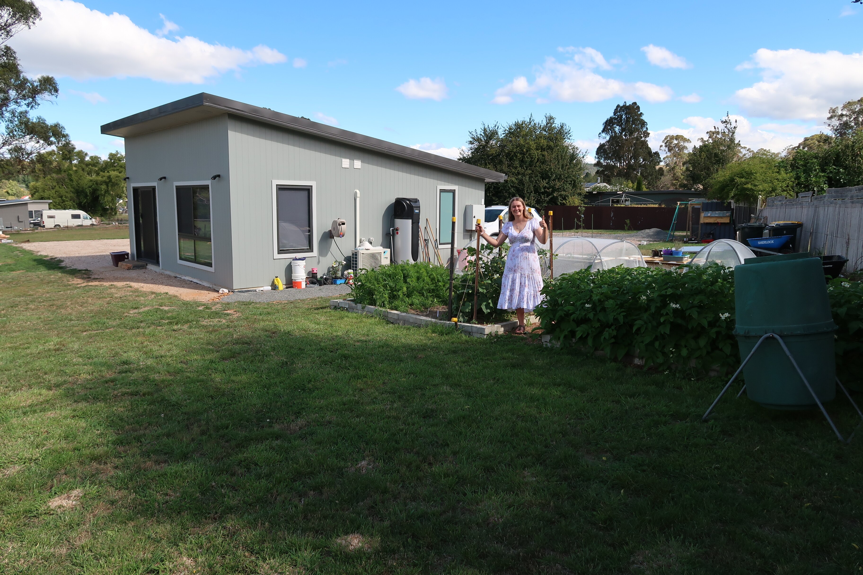 A young woman with long blond hair stands on a lawn next to a vegetable garden.