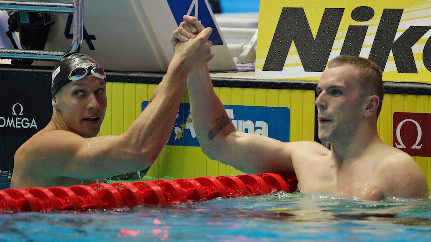Two male swimmers shake hands across a lane rope at the end of a race as they look at the scoreboard.