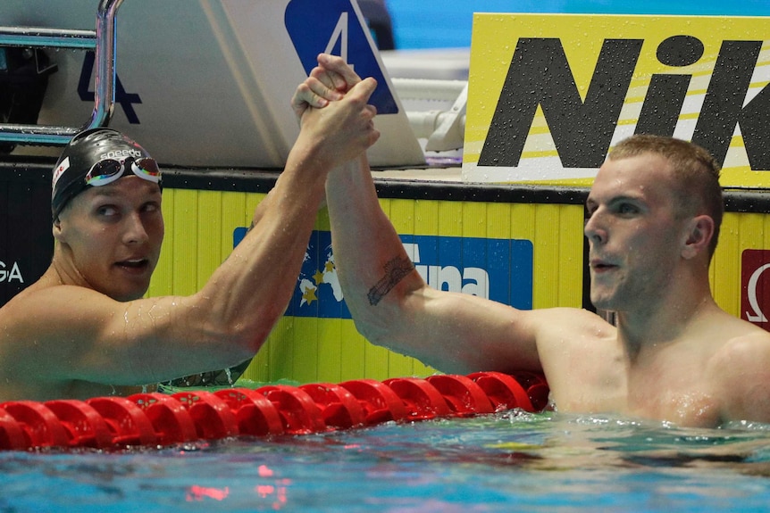 Two male swimmers shake hands across a lane rope at the end of a race as they look at the scoreboard.