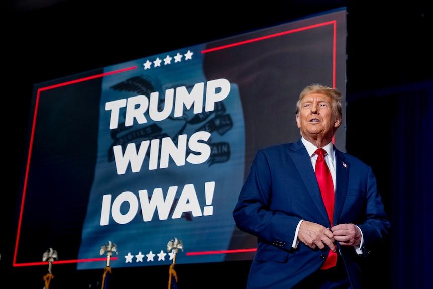Donald Trump, wearing a suit, appears next to the words 'Trump Wins Iowa' projected onto a screen