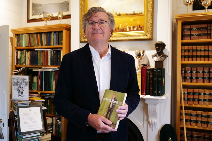 Andrew Leeming stands in his memorabilia room wearing a black blazer, white shirt and holding a cricket book.