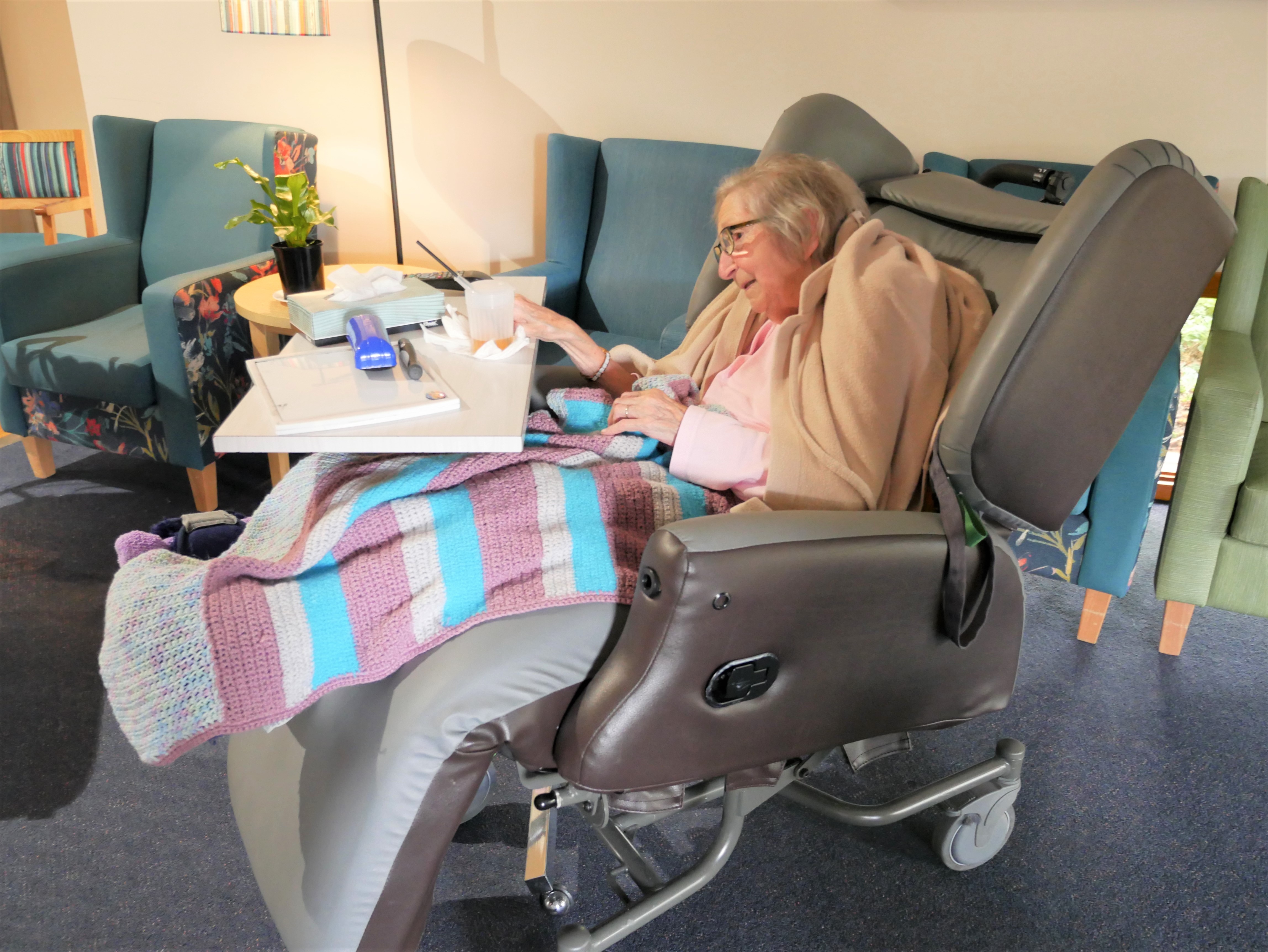 A woman sits in a special chair with a blanket over her legs and a food table.