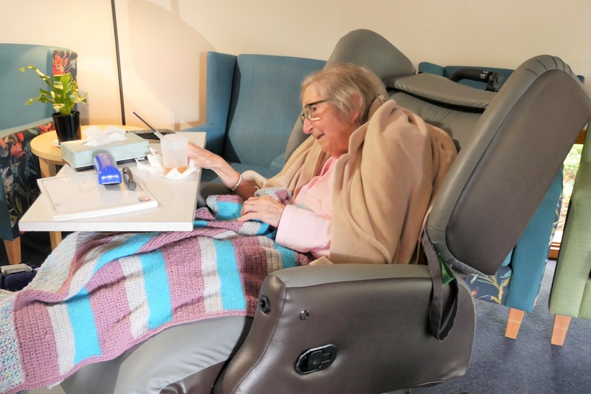 A woman sits in a special chair with a blanket over her legs and a food table.