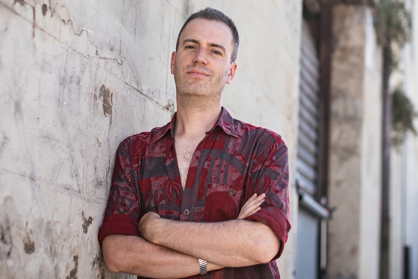 Man with short hair wearing dark red and black patterned shirt leaning against outdoor brick wall with street in background.