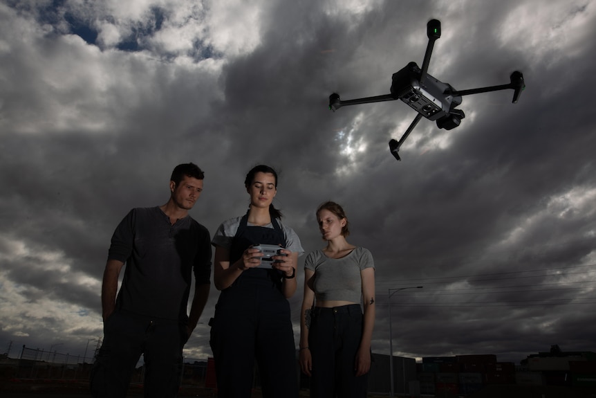 Two women and a man standing as the woman in the centre controls a drone camera.